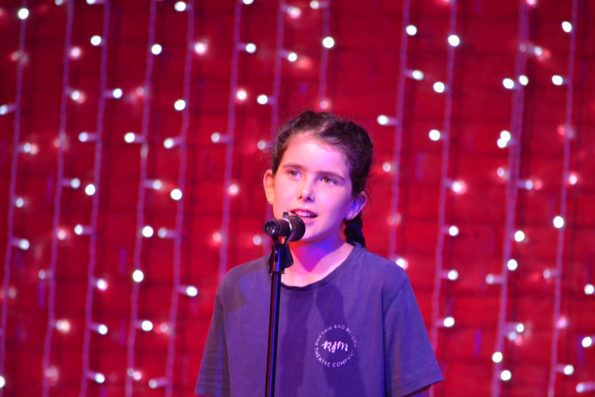 A performer sings at a microphone in front of a red curtain and fairy lights.