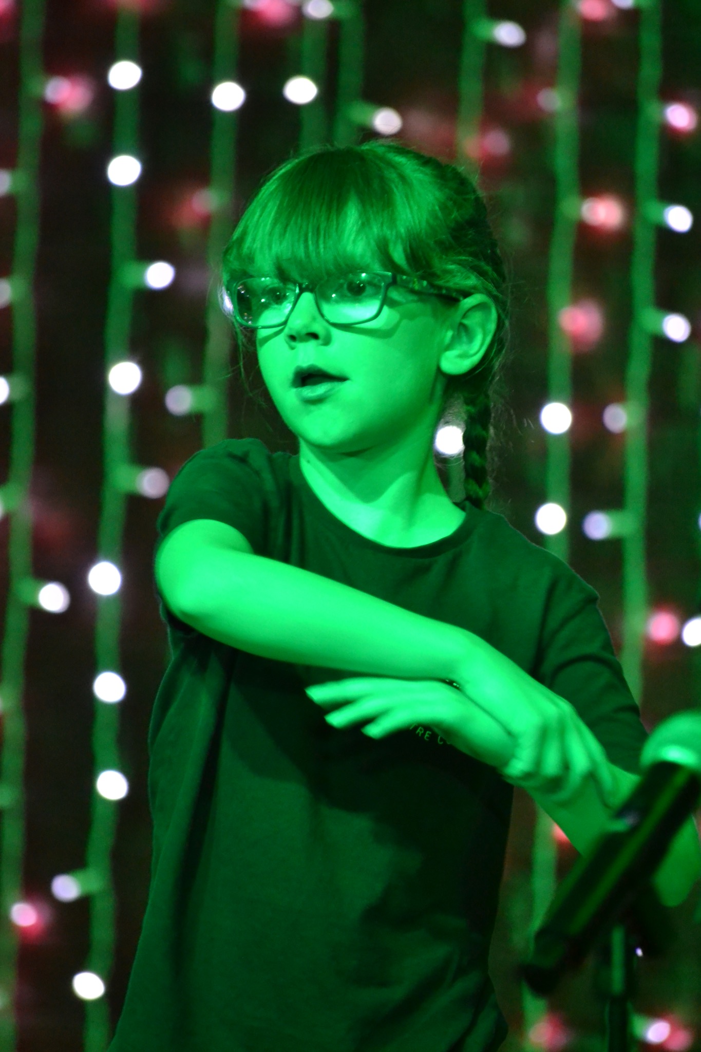 A performer wearing glasses gestures during a scene under green stage lighting.