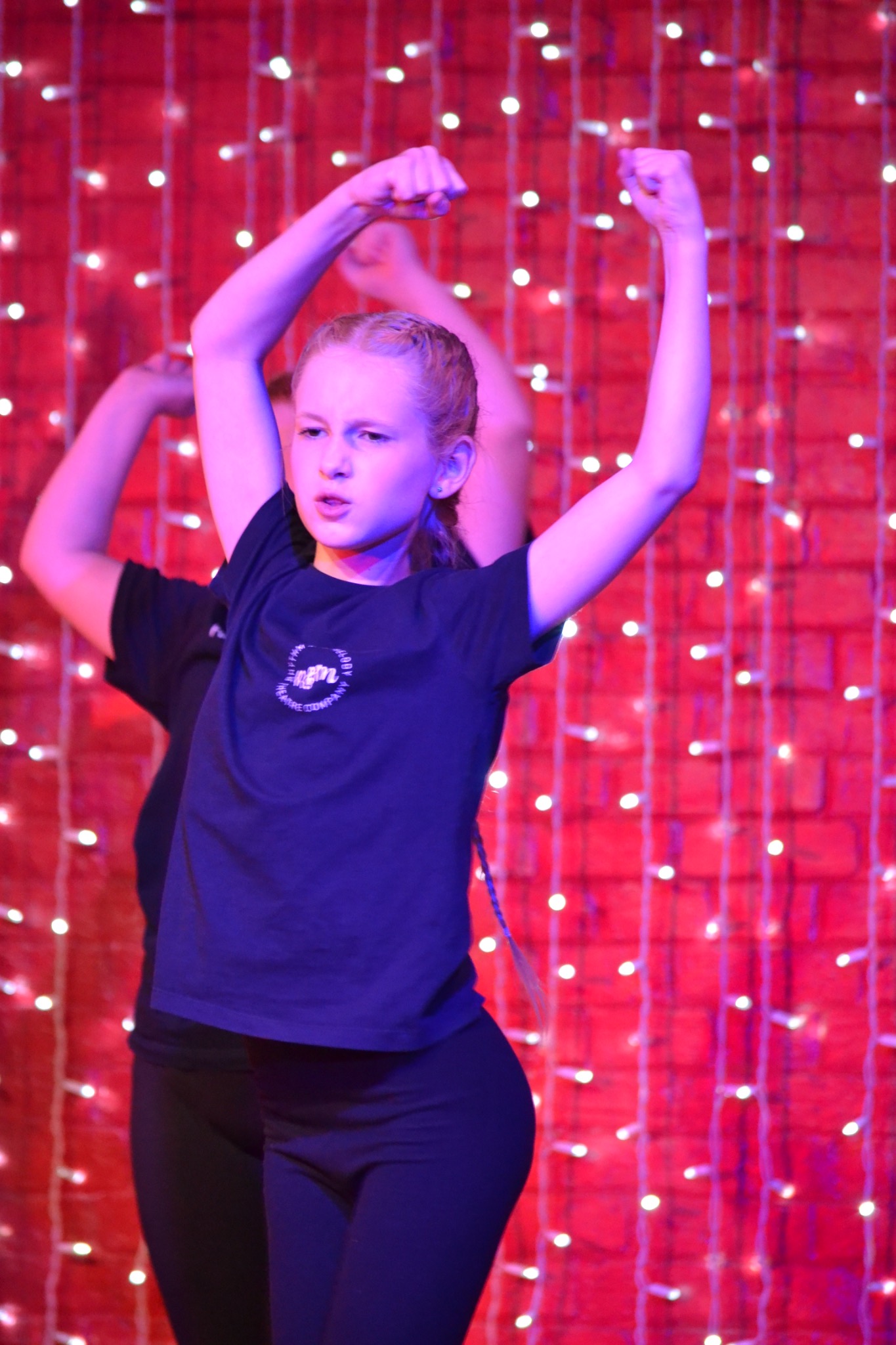 A performer raises both arms during a dance routine beneath colourful stage lights.